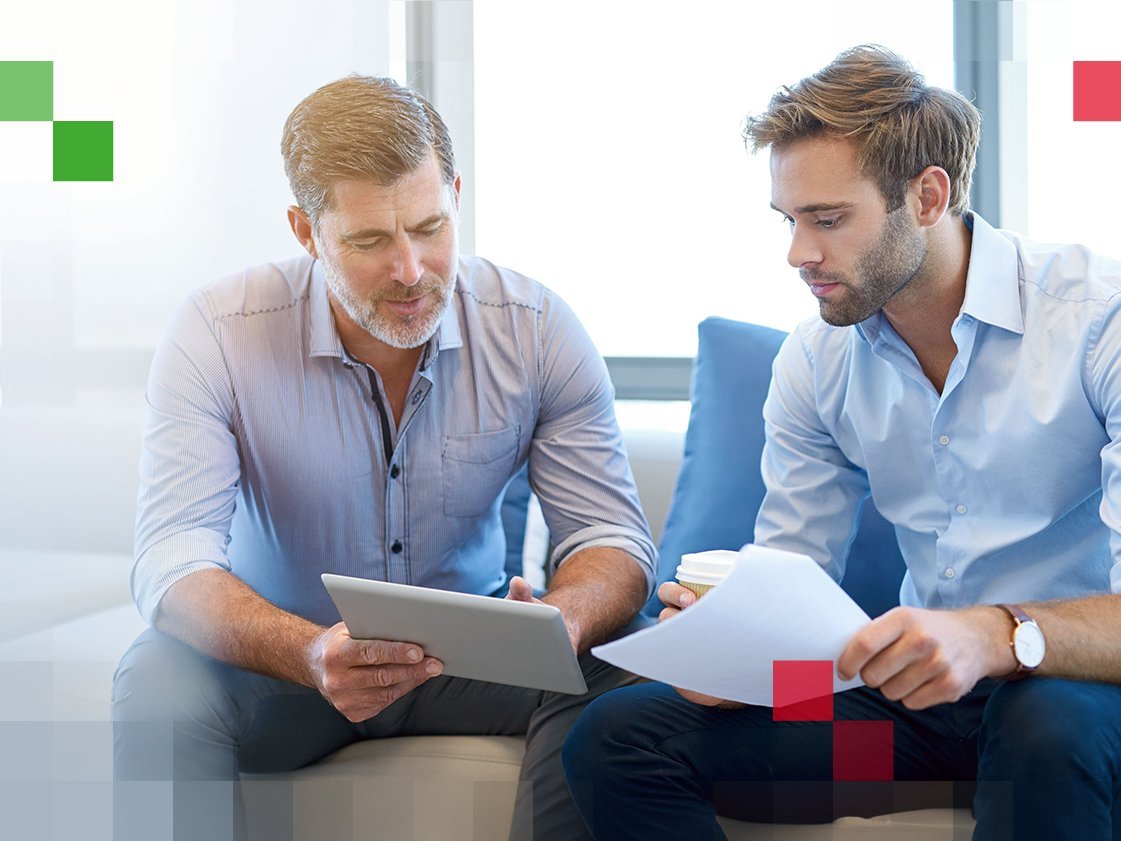 Zwei Männer in Hemd sitzen in einer Beratungssituation nebeneinander und deuten auf ein Tablet
